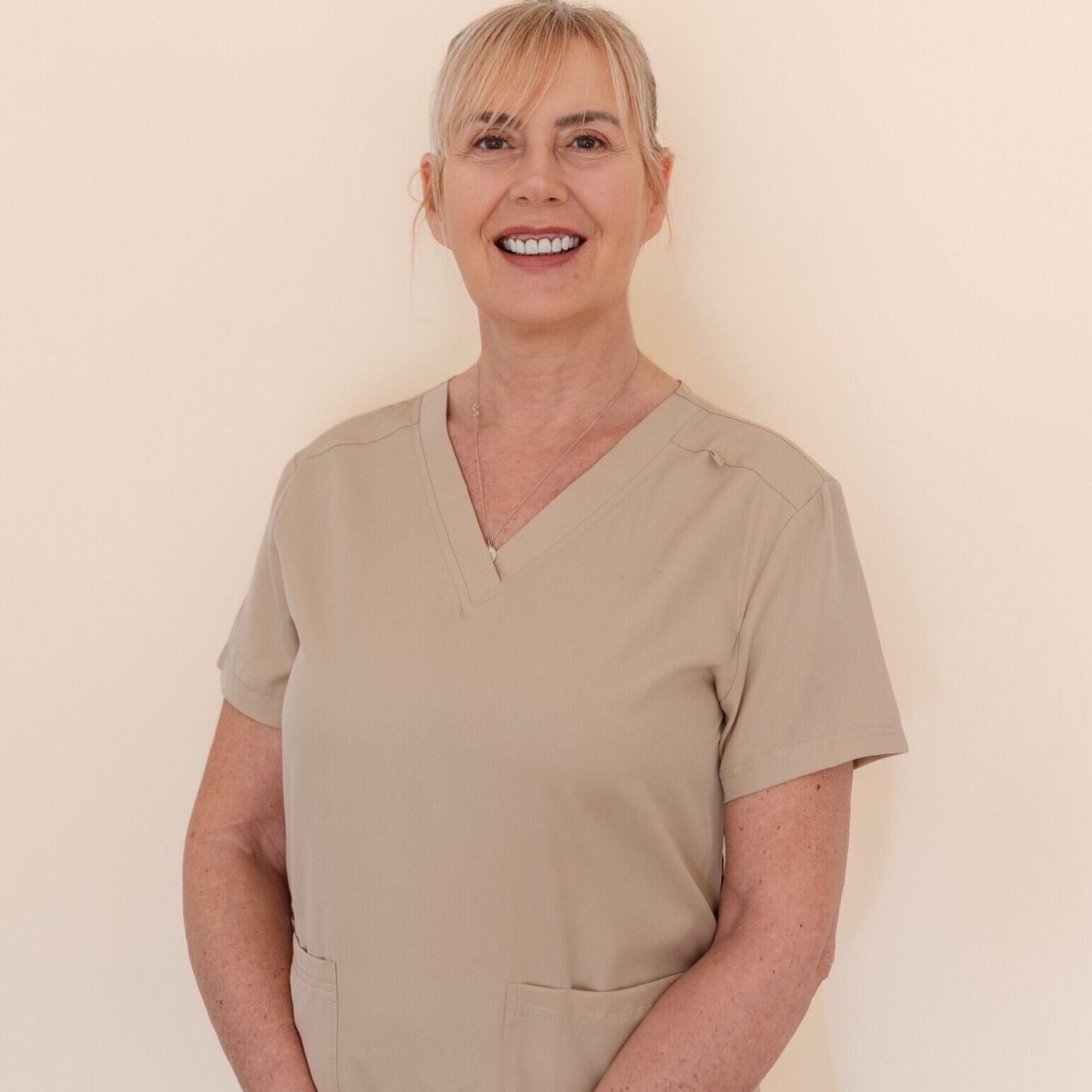 Smiling healthcare professional in beige scrubs stands against a plain background.